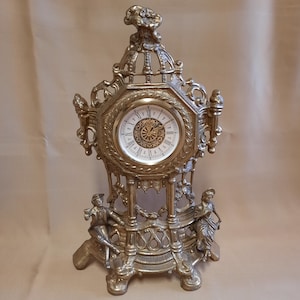 May include: A gold-toned, ornate clock with a white face and black Roman numeral hour markers. The clock is decorated with figures and intricate details, and it sits on a base with three legs.