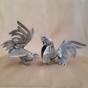 Può includere: Due figurine di gallo color argento con dettagli di piume e accenti rossi sugli occhi. Uno ha le ali spiegate, l'altro è in piedi con il becco aperto. Queste figurine decorative sono probabilmente per l'esposizione.
