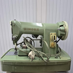 Pode incluir: Uma máquina de costura Singer vintage na cor verde sálvia com corpo metálico, um logótipo Singer proeminente na lateral, um painel de controlo com alavancas de ajuste verticais e um cabo de alimentação, montada numa base verde a condizer.