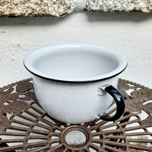 Op de afbeelding: Witte emaille kom met een zwarte rand en handvat. De kom heeft een gespikkeld patroon en staat op een decoratief metalen oppervlak.