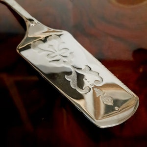Puede incluir: Un servidor de pastel de plata con un diseño grabado ornamentado. El servidor tiene una hoja larga y estrecha y un mango redondeado.