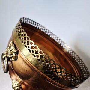 Könnte beinhalten: Eine kupferfarbene Schale mit Messingrand und Löwenkopfgriffen. Die Schale hat eine runde Form und steht auf drei Messingfüßen. Der Rand weist ein detailliertes, durchbrochenes Design auf. Die Schale ist wahrscheinlich vintage.