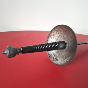 Op de afbeelding: Een schermfolie met een zwart, geribbeld handvat met een afgeronde pommel en een cirkelvormige, verweerde metalen handbeschermer met tekenen van oxidatie. Het dunne, metalen lemmet steekt naar voren uit het midden van de beschermer.