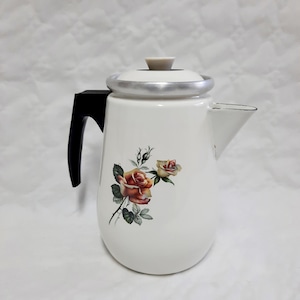 Op de afbeelding: Witte emaille koffiepot met een zwarte handgreep en een zilveren rand. De pot heeft een bloemenontwerp van oranje en gele rozen met groene bladeren. Het deksel heeft een kleine, beige knop.