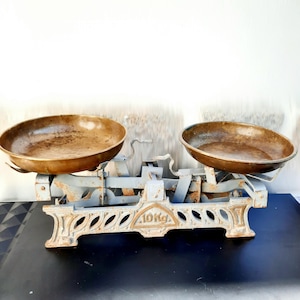 May include: An antique balance scale featuring two brass bowls and a distressed, pale blue painted metal frame. The scale is marked "10 Kg" and is designed for weighing items. The bowls show signs of age and use.