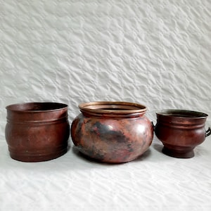 May include: Three copper pots of varying sizes. The largest pot has a rounded shape and a dark copper color with a reddish hue. The medium pot is a cylindrical shape with a smooth copper finish. The smallest pot is a rounded shape with a smooth copper finish.