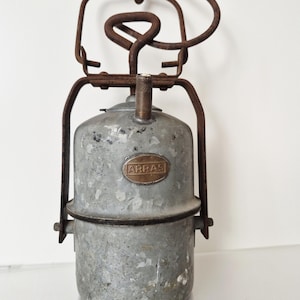 Op de afbeelding: Een antieke metalen lantaarn met een verweerde, gegalvaniseerde afwerking. De lantaarn heeft een cilindrisch lichaam, een handvat en een messing plaatje met het woord "ARRAS". Het frame en het handvat vertonen tekenen van roest, wat bijdraagt aan de vintage uitstraling.