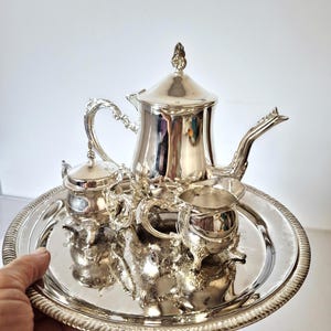 Op de afbeelding: Een gepolijste zilveren theeset bestaande uit een theepot met deksel, een suikerpot met deksel en een melkkannetje, gerangschikt op een rond zilveren dienblad met een decoratieve rand in touwstijl. Elk stuk heeft sierlijke handvatten.