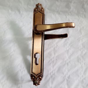 May include: A gold-toned door handle with a keyhole and ornate detailing. The handle has a traditional design with a curved lever and a rectangular plate.