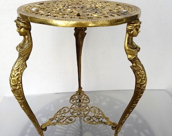 May include: A gold-colored, ornate plant stand with a round, openwork top. The stand features three legs, with two legs sculpted as figures and a central support. The stand is approximately 18 inches tall.
