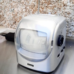 May include: Silver retro-style television with a clear, curved screen cover. The TV has a black dial and buttons on the side, and a retractable antenna. The brand name "DENVER" is printed on the front.
