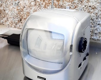 May include: Silver retro-style television with a clear, curved screen cover. The TV has a black dial and buttons on the side, and a retractable antenna. The brand name "DENVER" is printed on the front.