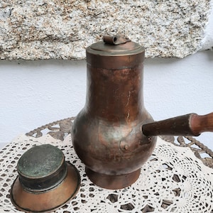 Könnte beinhalten: Eine antike Kaffeekanne aus Kupfer mit einem Holzgriff und einem Deckel. Die Kanne hat eine gehämmerte Oberfläche und ein Sieb.
