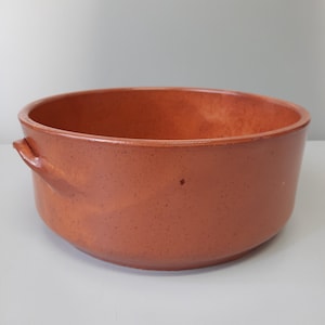 Puede incluir: Un cuenco redondo de cerámica color terracota con dos pequeñas asas a los lados. El cuenco tiene una textura moteada y un acabado liso y brillante. El interior es de un tono naranja ligeramente más claro. Este cuenco es adecuado para servir o exhibir.