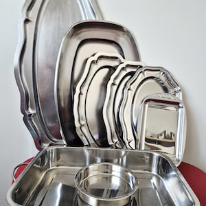 Peut inclure: Une collection de plateaux de service en acier inoxydable argenté de différentes formes et tailles, incluant des modèles ovales, rectangulaires et à bords festonnés, disposés avec un grand plat à rôtir rectangulaire et un petit bol rond.