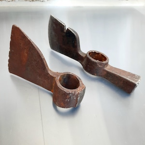 Op de afbeelding: Twee antieke, roestige metalen gereedschappen met een houweelachtig ontwerp. Elk gereedschap heeft een blad aan één kant en een cirkelvormige opening aan de andere kant, verbonden door een handvat. De gereedschappen worden waarschijnlijk gebruikt om te graven of materialen te breken.