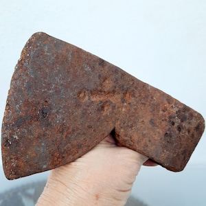 May include: A weathered axe head, showing signs of age and rust. The metal has a reddish-brown hue, with a textured surface. The blade is slightly curved. The axe head appears to be an antique tool, likely used for chopping wood. The overall length is approximately 20 cm.