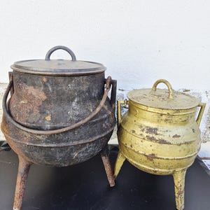 Puede incluir: Dos ollas antiguas de hierro fundido. La olla más grande es negra con un asa y una tapa. La olla más pequeña es dorada con una tapa y asas. Ambas ollas tienen tres patas y muestran signos de desgaste.