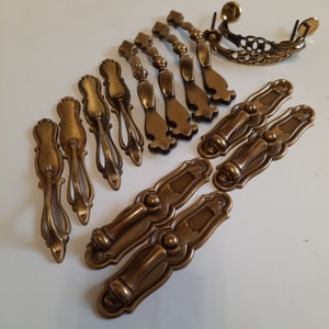 May include: A collection of 15 antique brass cabinet knobs and pulls. The knobs have a variety of styles, including ornate, simple, and modern. The pulls are all ornate and have a vintage look.