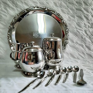 Puede incluir: Una bandeja de servir plateada con un borde decorativo, una tetera plateada, una jarra de crema plateada, un azucarero plateado y seis cucharas plateadas.
