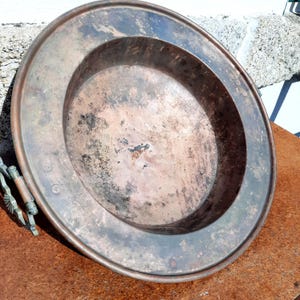 Puede incluir: Un plato grande de cobre antiguo con un borde ancho y un centro poco profundo. El plato tiene un aspecto deslustrado y envejecido con manchas oscuras y un asa en el lateral. Probablemente para servir o exhibir.