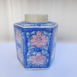 Pode incluir: Um pote de cerâmica hexagonal com um design floral azul e branco. O pote tem uma tampa e é decorado com flores cor-de-rosa e folhas verdes.