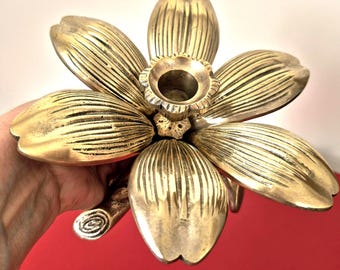 May include: A brass candle holder shaped like a six-petaled flower. The petals feature vertical ribbed detailing and a central circular cup for a candle. A small stem-like protrusion extends from the base of the flower-shaped metal structure.
