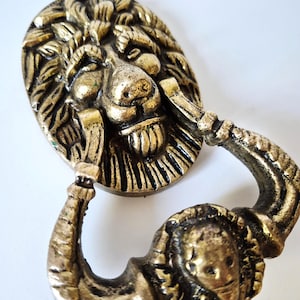 Op de afbeelding: Een messingkleurige metalen deurklopper met een gebeeldhouwde leeuwenkop op een ovale achterplaat. De zwaaiende hendel heeft de vorm van een gebogen arm met onderaan een klein, rond embleem van een leeuwengezicht met een verweerde afwerking.