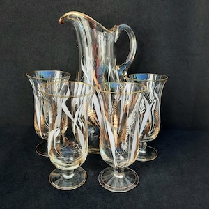 Puede incluir: Una jarra de cristal transparente y cuatro vasos a juego. La cristalería presenta un diseño de espiga de trigo en blanco y dorado. Los bordes de la jarra y los vasos están ribeteados en dorado. La jarra tiene un asa y un pico vertedor.