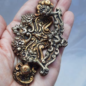 Op de afbeelding: Een decoratieve metalen deurgreep met een sierlijk ontwerp in goud- en zilverkleuren. De greep heeft ingewikkelde bloem- en krulpatronen.