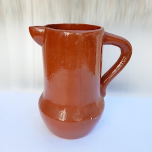 May include: A glazed, terracotta-colored pitcher with a rounded base and a curved handle. The pitcher has a spout for easy pouring. The ceramic jug is a rustic piece, perfect for serving beverages or as a decorative item.
