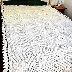 Puede incluir: Una colcha de ganchillo blanca hecha a mano con un patrón geométrico repetitivo de motivos cuadrados. Cada cuadrado está centrado con una aplicación floral tridimensional. Los bordes están rematados con un borde decorativo en forma de hojas.