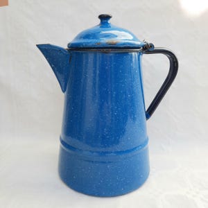 Op de afbeelding: Een vintage blauwe emaille koffiepot met een gespikkeld patroon. De pot heeft een deksel, een tuit en een zwarte handgreep. De buitenkant is helderblauw met witte spikkels, wat een klassieke camping- of keukenesthetiek oproept.