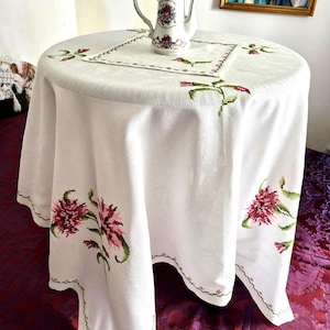 Pode incluir: Uma toalha de mesa redonda branca com padrões florais cor-de-rosa e caules verdes bordados em ponto cruz ao longo das bordas, acompanhada por um centro de mesa quadrado a condizer. Uma cafeteira de porcelana branca com tampa dourada.