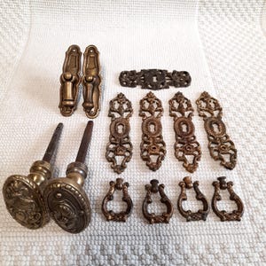 Puede incluir: Un conjunto de herrajes antiguos de latón, que incluyen pomos, bocallaves y tiradores. Las piezas presentan diseños ornamentados y una pátina cálida y envejecida. Los herrajes están dispuestos sobre una superficie blanca texturizada.