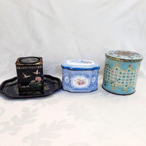 May include: Three vintage tin boxes with decorative designs. The first box is black with gold accents and a floral pattern. The second box is blue and white with a floral pattern. The third box is blue with a geometric pattern.
