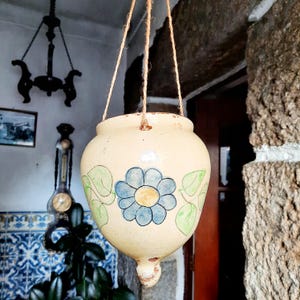 May include: Cream-colored hanging planter with a blue flower and green leaf design. The ceramic pot is suspended by twine and has a rounded shape with a small, pointed base. The background includes a glimpse of a rustic interior.