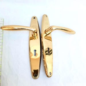 May include: A pair of vintage gold-coloured door handles. Each handle features a long, rectangular backplate with a keyhole and a curved lever. The handles are made of a shiny, reflective metal, possibly brass or a similar alloy. The handles are shown against a white background.