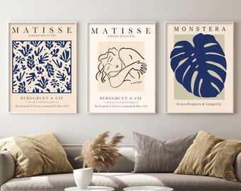 May include: Three minimalist art prints with a beige background and blue accents. The left print features a blue and white pattern of leaves and the text "MATISSE PAPIERS DECOUPES BERGGRUEN & CIE". The middle print features a line drawing of a woman in a blue outline and the text "MATISSE PAPIERS DECOUPES BERGGRUEN & CIE". The right print features a blue monstera leaf and the text "MONSTERA Honor, Respects & Longevity".