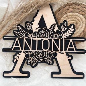 Könnte beinhalten: Ein Holzschild mit dem Buchstaben A und dem Namen Antonia in schwarzer Schrift, umgeben von einem floralen Design.