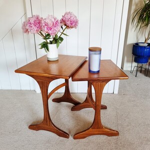May include: Two wooden nesting tables with a warm brown finish and curved, pedestal-style legs. A white ceramic vase filled with pink peony flowers sits on the larger table, while a blue glass candle with a wooden lid rests on the smaller table.