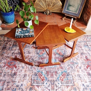 May include: A set of three brown wooden nesting tables with T-shaped legs. The largest table holds a potted Pilea plant and a book with a dark cover. The smallest table features a yellow ceramic candle holder with a white taper candle inside.