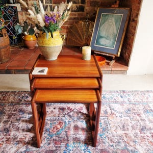 May include: A set of three nested tables crafted from medium-toned wood with a smooth finish. The tables feature rectangular tops and sturdy, straight legs, designed to slide neatly underneath one another for compact storage and versatile use.