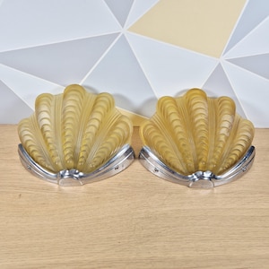 May include: A pair of wall lights featuring amber-coloured frosted glass shades shaped like seashells. Each shade is mounted on a polished silver-toned metal base with a curved, stepped design that follows the contour of the glass shell.