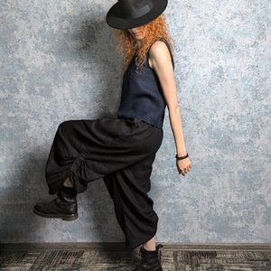 Op de afbeelding: Een vrouw draagt een zwarte fedora-hoed, een donkerblauw tanktop en een zwarte broek. Ze draagt zwarte laarzen en heeft rood krullend haar.