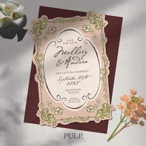 May include: A die-cut save the date card with a pale pink background and an ornate green floral border. The text reads: Save the Date, Mallory & Andrea are getting married, September 25th 2028, Provence, France, Formal invitation to follow.