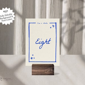 May include: A white card with a blue border and the text "Zoe + Martin" and "Eight" in blue cursive writing. The card is on a wooden stand.