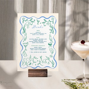 Könnte beinhalten: Eine Getränkekarte mit blauem Blumenrand und dem Wort "Drinks" in Schrift. Die Karte listet Bier, Wein und Cocktails auf. Rechts steht ein Cocktailglas mit weißem Schaum und einer dunkelroten Garnitur.