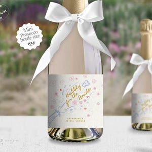 May include: A white mini prosecco bottle with a white satin ribbon tied around the neck. The bottle has a label with a hand-drawn illustration of a champagne bottle popping with the text "Bubbly for the Bride" and "Katherine's Bridal Shower".