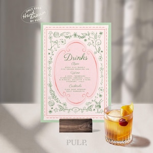 Può includere: Un menu di bevande rosa e verde con illustrazioni floreali e la parola "Drinks". Il menu è esposto su un supporto di legno, con un cocktail in un bicchiere in primo piano. Il menu elenca birre, vini e cocktail.
